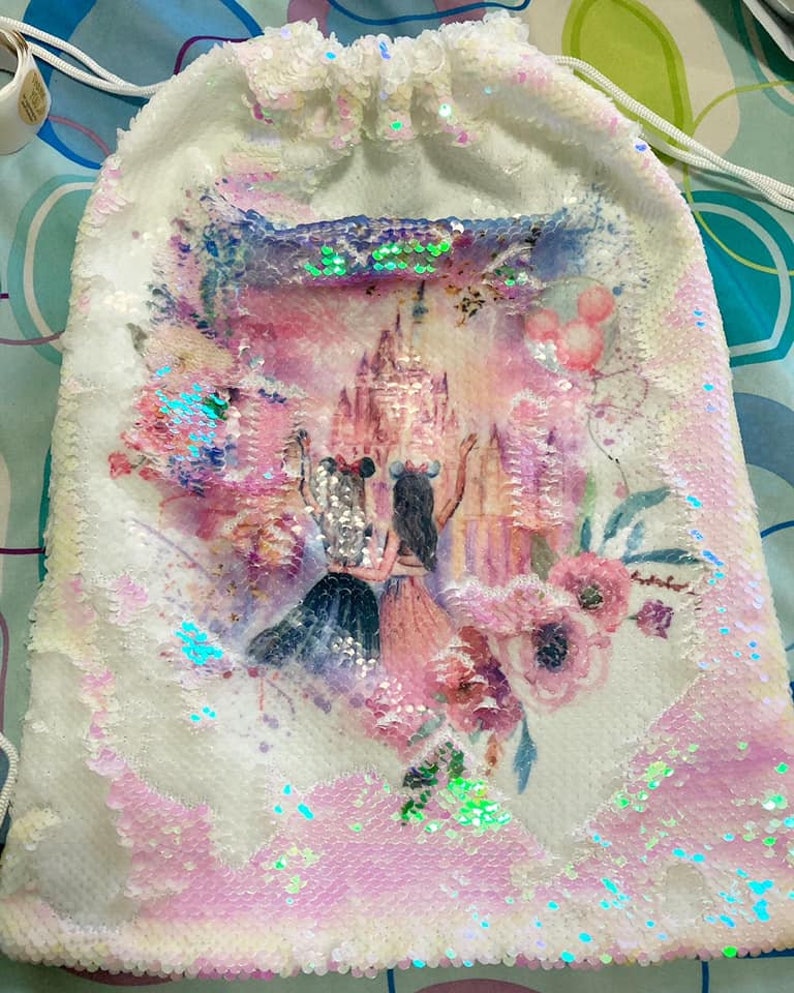 May include: White sequined drawstring bag featuring a colorful illustration of two figures in front of a castle, with pink and purple floral accents. The bag has a white drawstring closure and is covered in iridescent sequins.