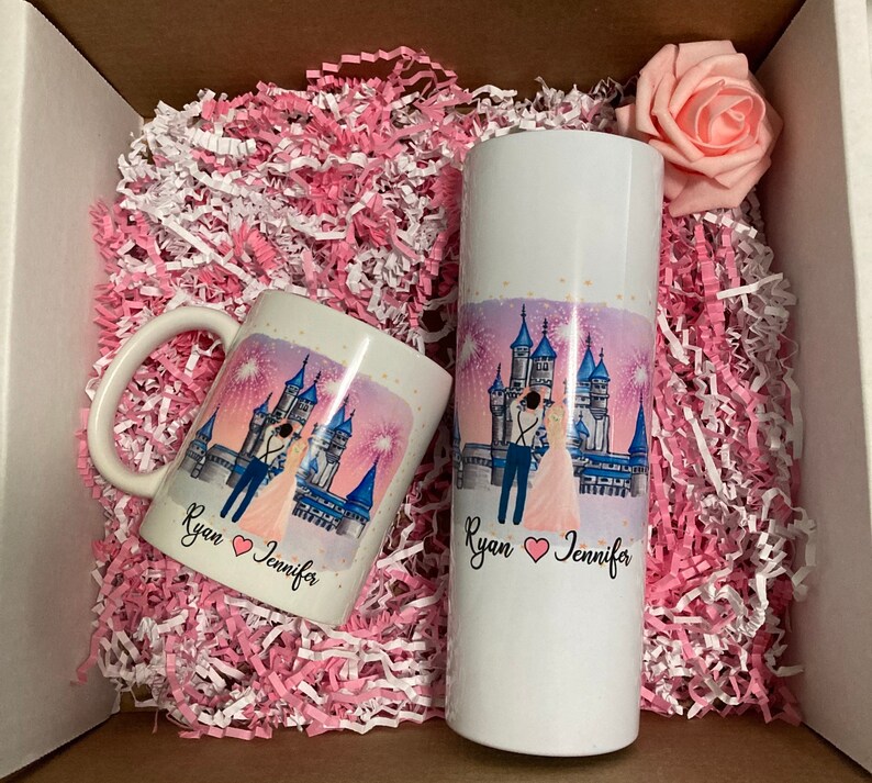 May include: A white mug and a white tumbler with a pink and blue castle illustration and the text "Ryan Jennifer" with a heart symbol between the names. The mug and tumbler are in a white box with pink and white shredded paper.