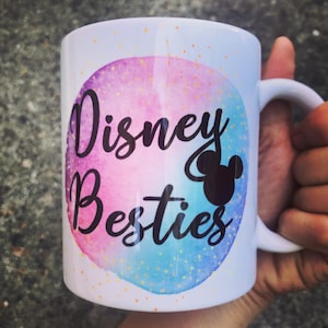 May include: White ceramic mug with the words "Disney Besties" in black script, set against a watercolor background of pink and blue. A black silhouette of Mickey Mouse is also featured. The mug has a white handle.
