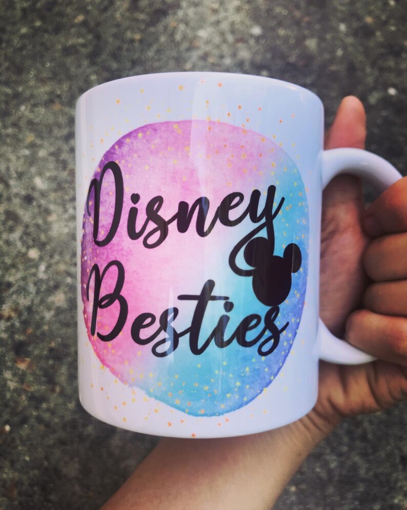 May include: White ceramic mug with the words "Disney Besties" in black script, set against a watercolor background of pink and blue. A black silhouette of Mickey Mouse is also featured. The mug has a white handle.
