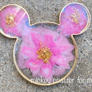 Pu&ograve; includere: Sottobicchiere in resina rosa e bianca a forma di orecchie di Topolino con glitter e scaglie dorate. Il sottobicchiere ha il testo "mickey coaster for mug" in basso.