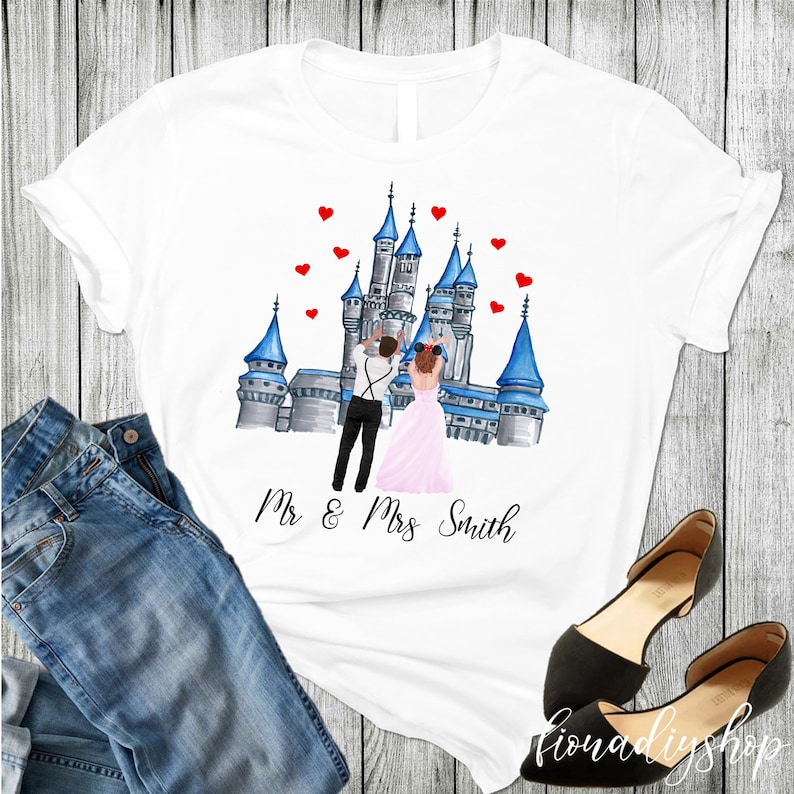 May include: White t-shirt with a watercolor illustration of a couple in front of a castle. The castle is blue and gray with red hearts floating above it. The text "Mr & Mrs Smith" is printed below the illustration.