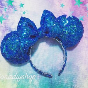 May include: Blue sequin Minnie Mouse ears with a large blue bow. The ears are on a white background with a light blue and purple watercolor effect.