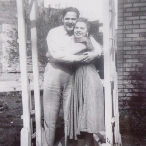May include: A black and white photo of a couple embracing in front of a wooden trellis. The man is wearing a button-down shirt and the woman is wearing a dress.