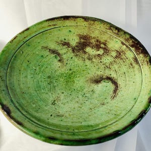 Peut inclure: Une assiette ronde en céramique présentant un émail vert clair moucheté avec des accents et des stries brun foncé, couleur rouille, sur toute la surface. L'assiette possède un large bord plat et un centre légèrement creusé, à l'aspect texturé.