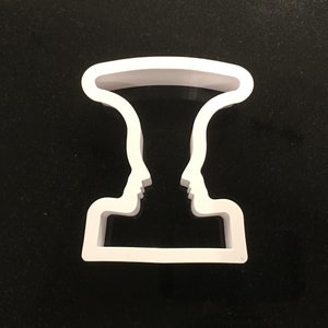 May include: White plastic cookie cutter shaped like two profiles facing each other, forming a vase shape.
