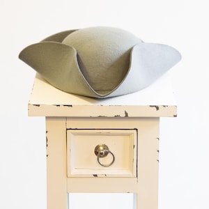 Peut inclure: Un chapeau tricorne en feutre gris clair posé sur une table d'appoint en bois blanc vieilli avec un seul tiroir. Le tiroir a une poignée argentée.