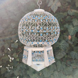 May include: A white birdcage with a blue and white wire design. The cage is round with a square base and has a hook on top.