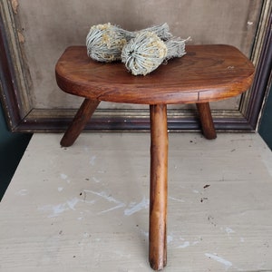 May include: A rustic wooden stool with three legs. The stool is made of dark brown wood and has a rounded top. The stool is sitting on a light brown surface.