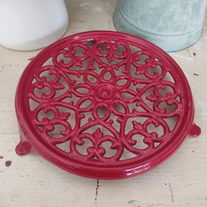 May include: Red cast iron trivet with a decorative, intricate design. The trivet has a round shape and three feet.