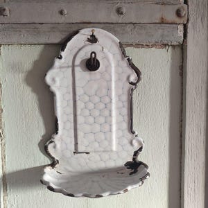 May include: Antique white ceramic wall-mounted soap dish with a decorative, ornate design. The dish features a textured, honeycomb-like pattern and a small shelf. The edges are trimmed with a dark, aged finish, and it has a metal hanging loop.