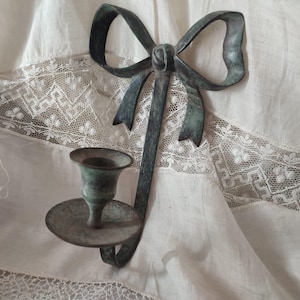 Può includere: Un portacandele da parete in stile vintage con un design a fiocco. Il portacandele in metallo ha una finitura anticata, verde rame. Il portacandele è montato su un tessuto bianco con finiture in pizzo.