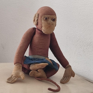 Peut inclure: Un singe en peluche vintage avec une fourrure brune, une jupe en tissu bleu et une longue queue brune. Le singe a un visage cousu avec des yeux bruns et une bouche fermée.