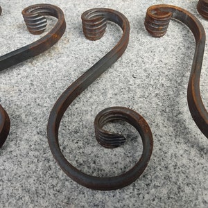 Może przedstawiać: Cztery rustykalne metalowe wzory zwoju z misternymi detalami. Zwoje są ułożone w wzór na szarej, fakturowanej powierzchni.