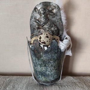 Peut inclure: Un petit seau en métal gris avec une bordure en dentelle blanche et un dessus en forme de lapin argenté. Le seau est rempli d'œufs de caille tachetés.