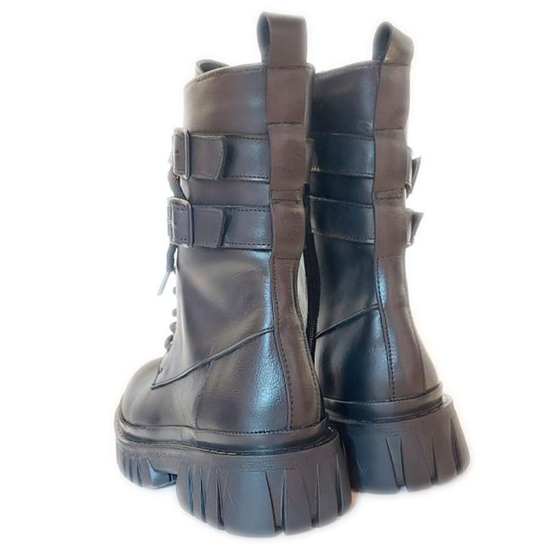 K&ouml;nnte beinhalten: Ein Paar schwarze Leder-Combat-Stiefel mit klobiger Sohle. Die Stiefel verf&uuml;gen &uuml;ber drei Schnallenriemen und ein Schn&uuml;rdesign. Die Stiefel haben eine Zuglasche oben und sind vor einem wei&szlig;en Hintergrund platziert.