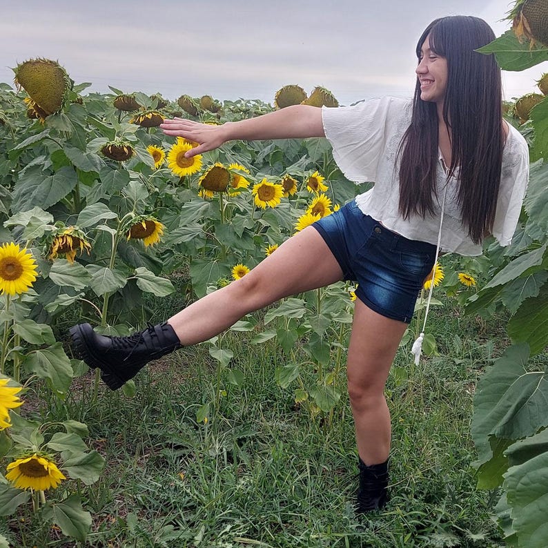 K&ouml;nnte beinhalten: Eine Person in einem Sonnenblumenfeld, bekleidet mit einem wei&szlig;en Oberteil, Jeansshorts und schwarzen Stiefeln. Die Sonnenblumen befinden sich in verschiedenen Bl&uuml;hstadien, mit leuchtend gelben Bl&uuml;tenbl&auml;ttern und dunkelbraunen Zentren. Der Himmel ist bew&ouml;lkt.