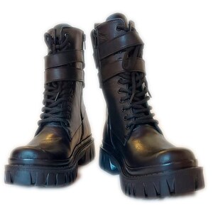 K&ouml;nnte beinhalten: Ein Paar schwarze Leder-Combat-Stiefel mit dicken Sohlen. Die Stiefel haben Schn&uuml;rsenkel, seitliche Rei&szlig;verschl&uuml;sse und verstellbare Riemen mit Schnallen. Die hoch geschnittenen Stiefel haben eine Plateausohle und bieten einen k&uuml;hnen, kantigen Look.