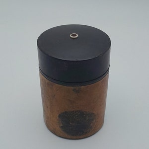 May include: A round wooden container with a black lid and a small hole in the centre. The container has a dark brown stain on the side.
