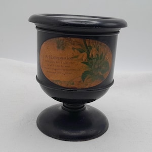 May include: A black keepsake urn with a pedestal base and a wide rim. The urn has an oval-shaped label with floral imagery and the text "A Keepsake". The urn is made of a dark, possibly painted, material.