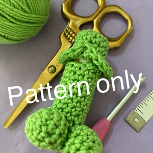 Könnte beinhalten: Eine grüne gehäkelte Amigurumi-Figur, die der menschlichen männlichen Anatomie ähnelt. Die Figur liegt auf einer violetten Oberfläche mit einer goldenen Schere, einem rosa Häkelhaken und einem Lineal.