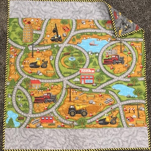 May include: A colorful, quilted play mat featuring a construction site design with roads, buildings, and construction vehicles. The mat includes the text "Tonka" and is bordered with a black and yellow hazard stripe.