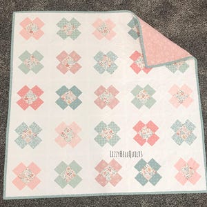 May include: A handmade quilt featuring a repeating floral pattern in pink, blue, and white. The quilt has a white background with a floral pattern in the center of each square. The quilt is reversible with a pink floral pattern on the back. The text "LizzyBellQuilts" is visible.