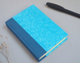 Op de afbeelding: Een klein blauw notitieboekje met een marmeren omslag en een donkerblauwe rug. Het notitieboekje is gesloten en ligt op een lichtgrijze ondergrond.