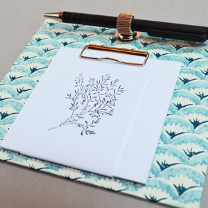 Op de afbeelding: Een blauw en wit klembord met een bloemenpatroon en een bevestigde pen. Een wit vel papier is met een tekening van een plant aan het klembord geklikt.