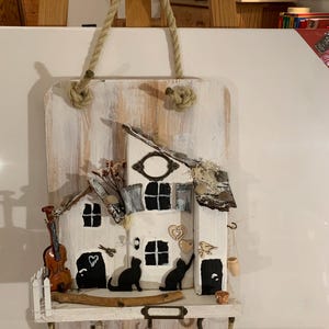 Könnte beinhalten: Ein rustikaler, weißer Holz-Schlüsselhalter mit hausförmigem Design. Mit einer Miniatur-Geige, schwarzen Katzen-Silhouetten und einem kleinen Vogel. Enthält einen Seilgriff und dekorative Elemente.