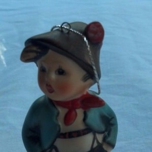 May include: A ceramic figurine of a young boy wearing a brown hat with a red pom-pom, a teal jacket, and black shorts. The boy has rosy cheeks and is standing on a round, beige base. A silver string is attached to the hat.