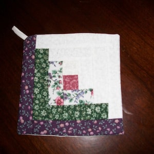 May include: Square quilted potholder with a white center and a log cabin design. The design incorporates green, floral, and purple patterned fabrics. A white loop is attached for hanging.