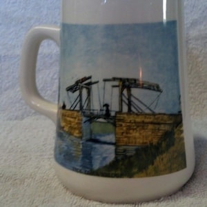 May include: White ceramic mug with a handle, featuring a painting of a bridge over water. The artwork depicts a bridge with a raised section, a person, and a blue sky. The mug has a curved shape.