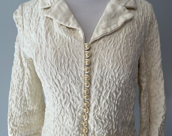 May include: A white, textured, cropped jacket with a collar and long sleeves. The jacket has a button closure down the front with pearl-like buttons.