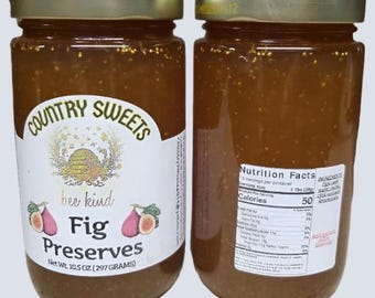 May include: Two glass jars of Country Sweets fig preserves with gold metal lids. The front label features a beehive illustration, the text 'bee kind', and 'Fig Preserves', with a net weight of 297 grams. The back label displays a nutrition facts table.