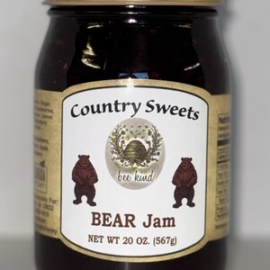 May include: A glass jar of dark fruit jam with a gold metal lid. The label features the text Country Sweets, an illustration of a beehive, two brown bears, and the text bee kind and BEAR Jam. The label states a net weight of 20 oz. (567g).