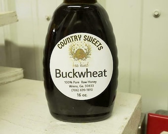 May include: A dark glass bottle of buckwheat honey with a yellow cap. The label reads "Country Sweets" and "Buckwheat." The bottle contains 16 oz of honey.