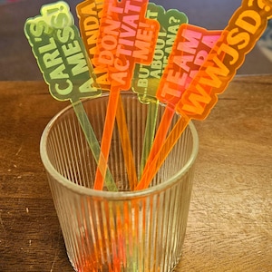 May include: A clear glass tumbler holds several colorful drink stirrers. The stirrers are orange and green with text that reads "CARL'S A MESS!", "AMANDA DON'T ACTIVATE", "BUT ME!", "TEAM ADA ABOUT ANA?", and "WWJSD?".