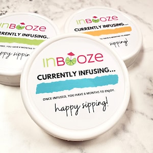 Puede incluir: Tres recipientes redondos blancos con tapas. Cada tapa tiene una etiqueta colorida con el texto "inBooze" y la frase "Currently Infusing..."  Las etiquetas tambi&eacute;n incluyen una lima y un paraguas de playa. Una etiqueta tiene el texto "Una vez infundido, tienes 6 meses para disfrutarlo. &iexcl;Feliz degustaci&oacute;n!"