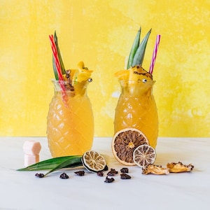 May include: Two pineapple-shaped glasses filled with a yellow drink, garnished with pineapple, starfruit, and lime slices. The glasses are on a white surface with dried fruit slices and pink sugar cubes.