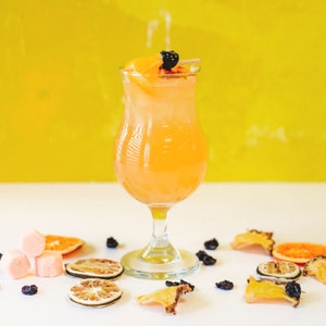 May include: A tall glass filled with a light orange drink garnished with a slice of orange and a black berry. The glass is on a white surface with dried fruit slices and black berries scattered around it.