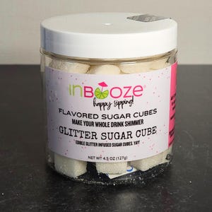 May include: A clear glass jar filled with white sugar cubes that have edible glitter infused in them. The jar has a white lid and a pink and white label that reads "inBoze happy sipping!" "FLAVORED SUGAR CUBES MAKE YOUR WHOLE DRINK SHIMMER GLITTER SUGAR CUBE EDIBLE GLITTER INFUSED SUGAR CUBES, YAY! NET WT 4.5 OZ (127g)"