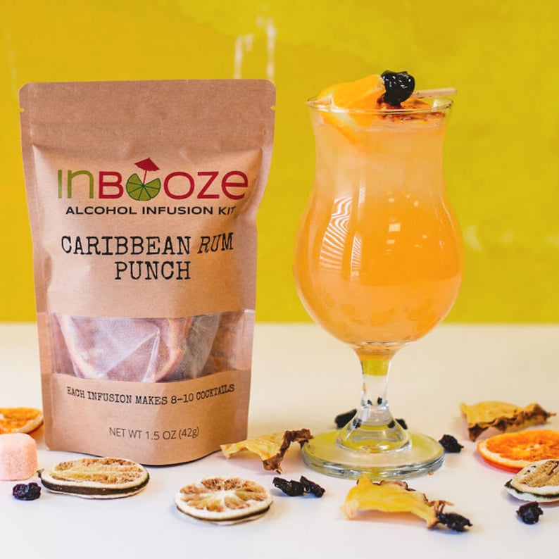 May include: A brown paper pouch with the text "InBooze Alcohol Infusion Kit Caribbean Rum Punch" and the text "Each infusion makes 8-10 cocktails Net Wt 1.5 oz (42g)". A glass of orange punch with a slice of orange and a black berry on top. The glass is sitting on a white surface with dried fruit slices and black berries.