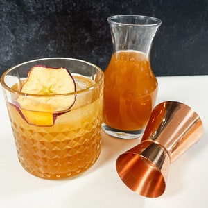 May include: A close-up of a cocktail with an apple slice garnish in a clear glass with a diamond pattern. A copper jigger and a clear glass decanter with a brown liquid are also in the image.