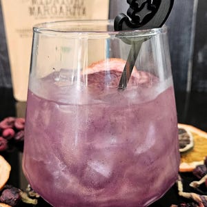 May include: A clear glass filled with a purple liquid and ice cubes, featuring a black drink stirrer shaped like a crescent moon with stars. The glass is positioned in front of a tan package labeled Midnight Margarita with dried fruit slices.