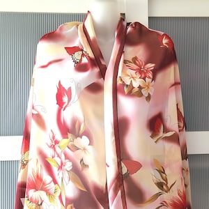 Op de afbeelding: Een doorschijnende sjaal met een bloemen- en vlinderpatroon. De sjaal heeft rode, witte en gele bloemen en rode vlinders tegen een crème- en bordeauxrode achtergrond. De sjaal is over een mannequin gedrapeerd.