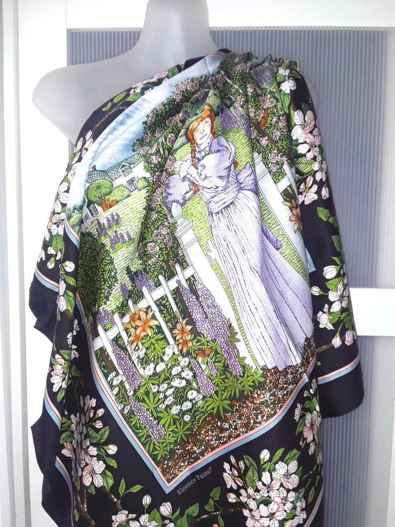 May include: A black silk scarf with a colorful floral print featuring a woman standing in a garden with a white picket fence. The scarf has a blue border and the text "Eleanor Paine" in the bottom right corner.