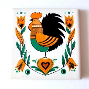 May include: A white ceramic tile featuring a colourful rooster design. The rooster is orange, green and black with a red comb. The tile is surrounded by green leaves and orange flowers.