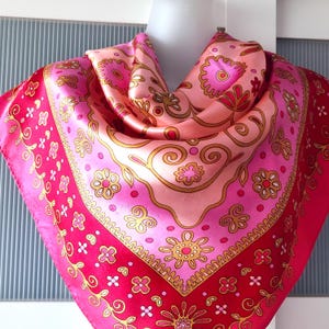 May include: A square silk scarf in pink and red hues, featuring gold floral and swirl designs. The scarf is displayed on a mannequin head, highlighting its pattern and texture. The borders are a deeper red, contrasting with the lighter pink central area.