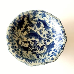 May include: A white ceramic bowl with a blue and white design of fish swimming in water. The bowl has a scalloped edge and is decorated with a floral pattern.